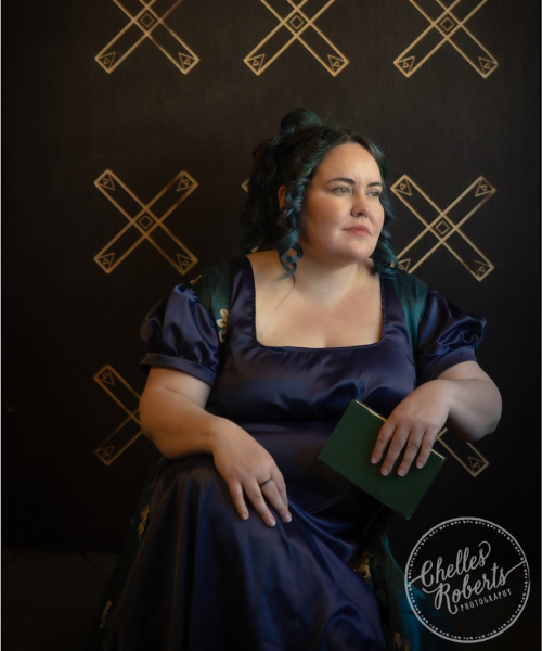 Signature Image Frances is female presenting, dressed in full regency attire, a blue dress and patterned green robe. Dark brown, blue and green hair falls in ringlets, framing the face. In the left hand is an old green book, on the right middle finger is a black ring. Frances is looking to the right into the light. In the rear is a piece of art.